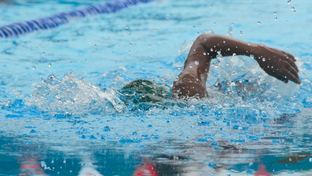 A swimmer's arm and shoulder breaking the surface of a pool lane, water droplets bright on the skin