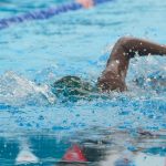 A swimmer's arm and shoulder breaking the surface of a pool lane, water droplets bright on the skin