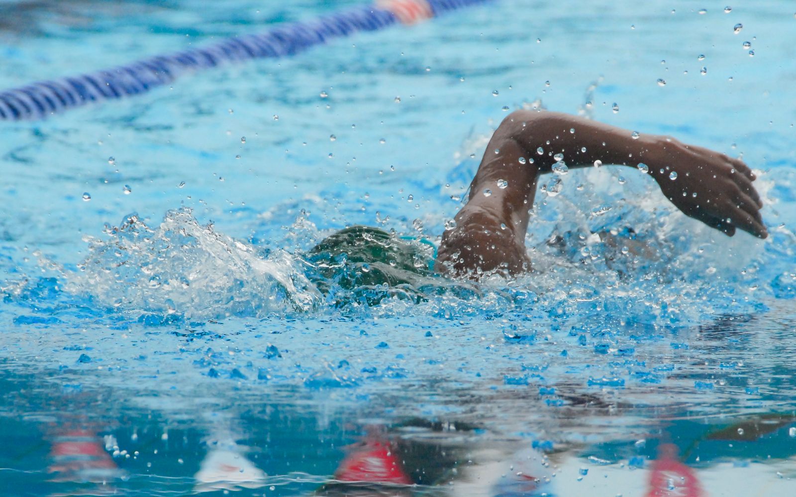 A swimmer's arm and shoulder breaking the surface of a pool lane, water droplets bright on the skin