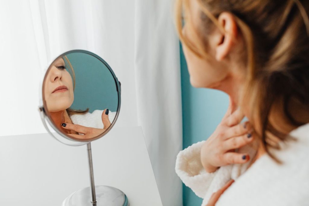A woman in a white bathrobe smoothing moisturizer along her neck with her fingertips while checking a small round mirror, soft daylight