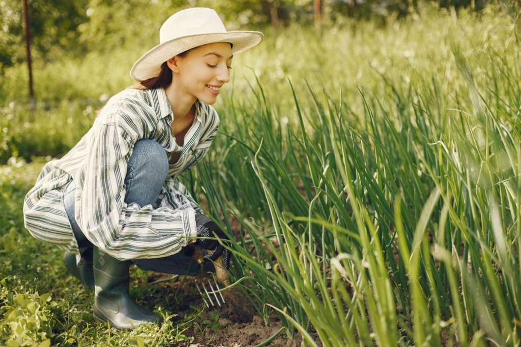 A woman in a sunhat and striped shirt kneeling in a sunny garden among tall grasses