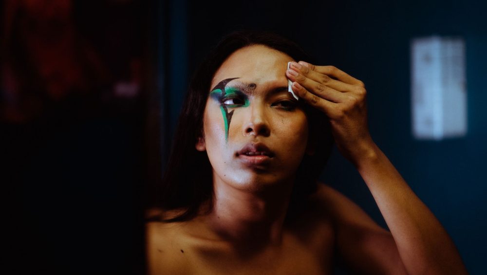 A woman wiping away dramatic eye makeup near her brow with a small folded pad, low evening light