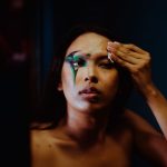 A woman wiping away dramatic eye makeup near her brow with a small folded pad, low evening light
