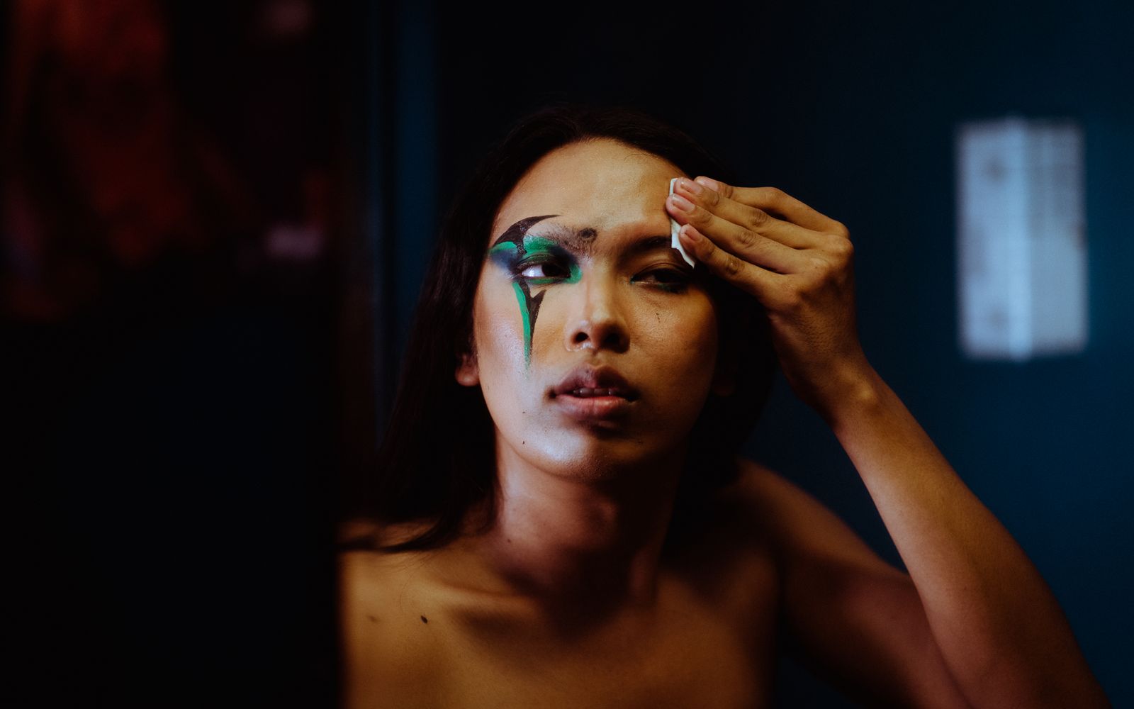 A woman wiping away dramatic eye makeup near her brow with a small folded pad, low evening light