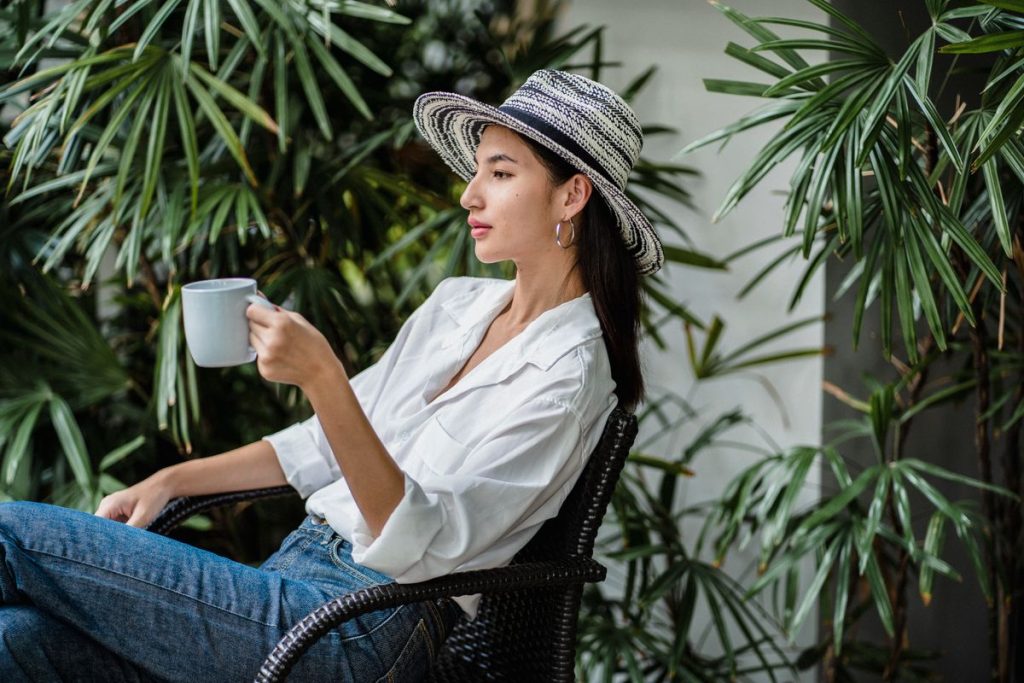 A woman in a white long-sleeved shirt and a sunhat sitting outdoors among plants with a morning drink