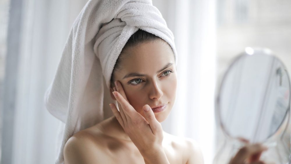 A woman with a towel wrapped around her hair touching her cheek with her fingertips while holding a hand mirror, soft window light