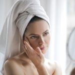 A woman with a towel wrapped around her hair touching her cheek with her fingertips while holding a hand mirror, soft window light