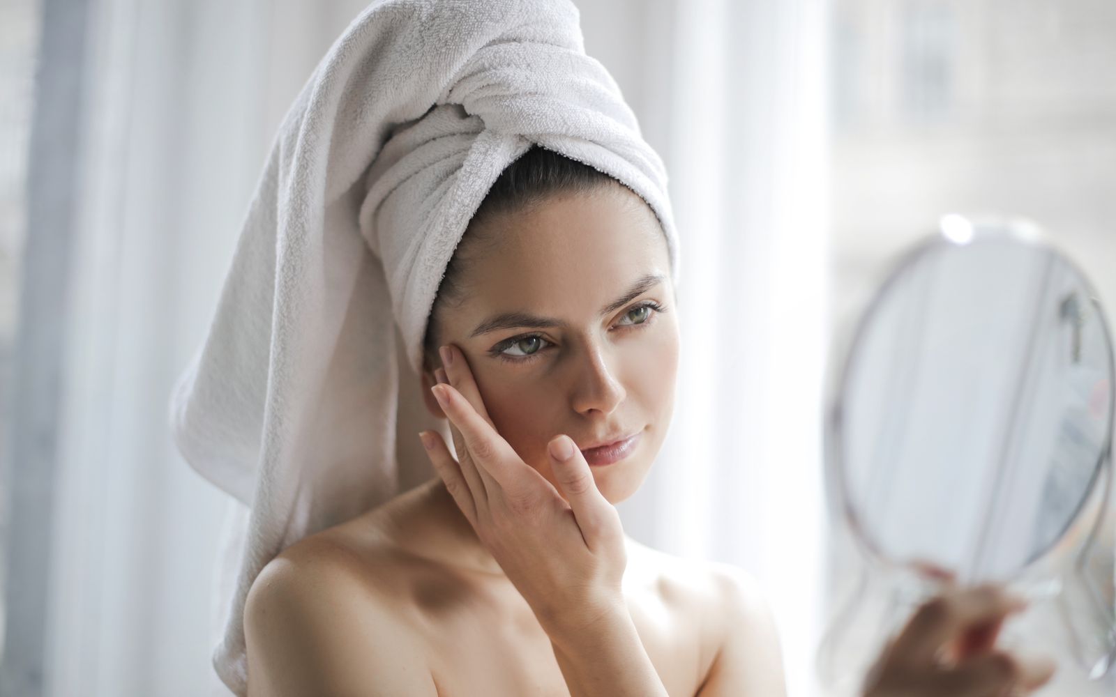 A woman with a towel wrapped around her hair touching her cheek with her fingertips while holding a hand mirror, soft window light