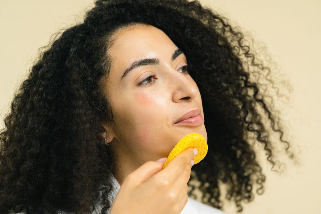 A woman gently pressing a soft yellow facial sponge against her jaw, plain warm background