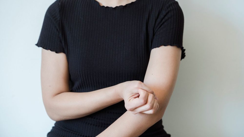 A woman in a dark T-shirt holding her forearm against a plain light background