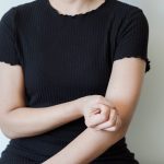 A woman in a dark T-shirt holding her forearm against a plain light background